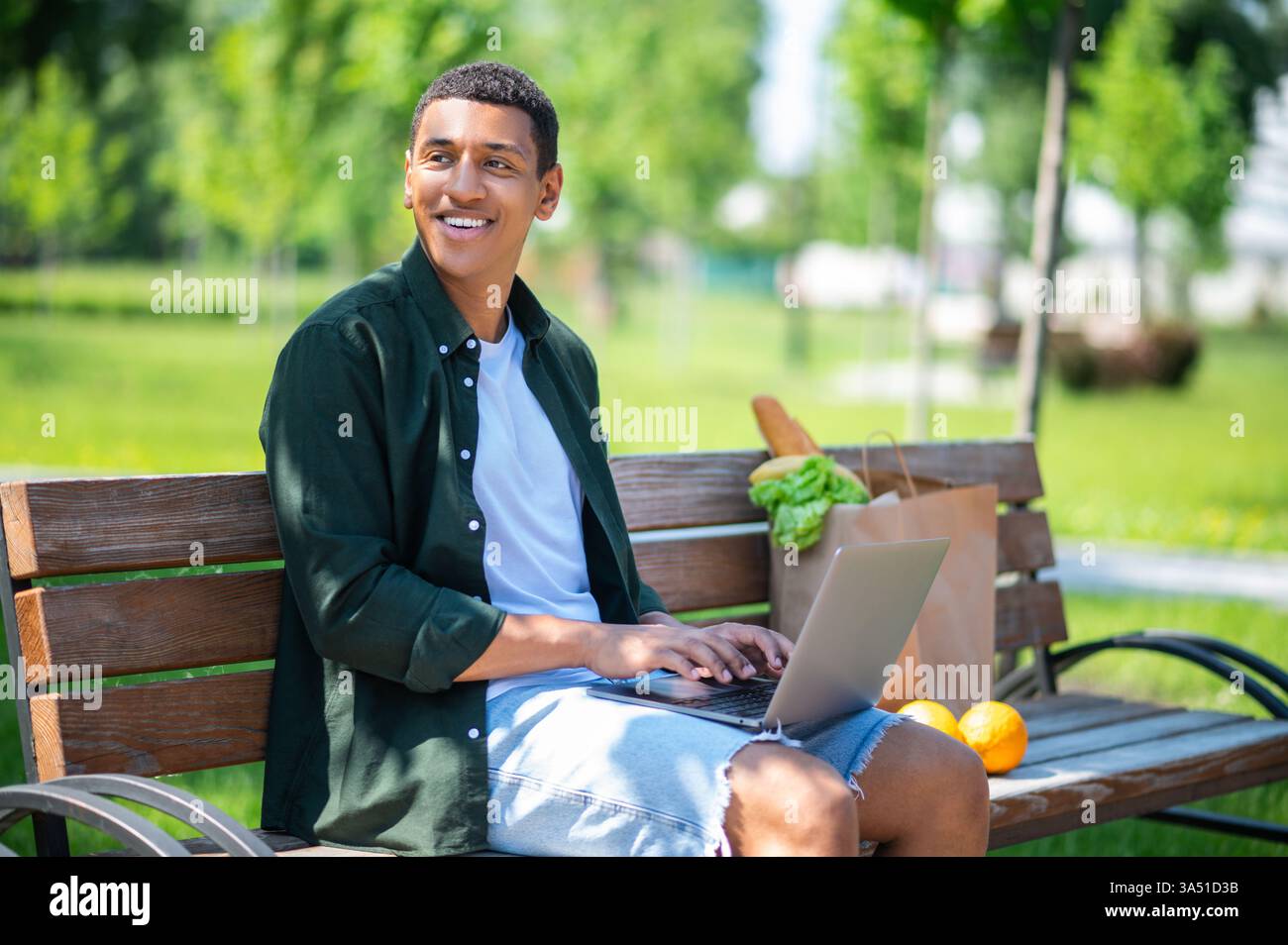 Un homme noir joyeux est assis sur un banc de parc, travaillant sur son ordinateur portable avec un sac de collations à proximité par une journée ensoleillée. Idéal pour le travail à distance, les loisirs en plein air et les thèmes de style de vie sain dans les parcs. La scène transmet la concentration détendue, l'énergie positive et la vie urbaine décontractée. Banque D'Images