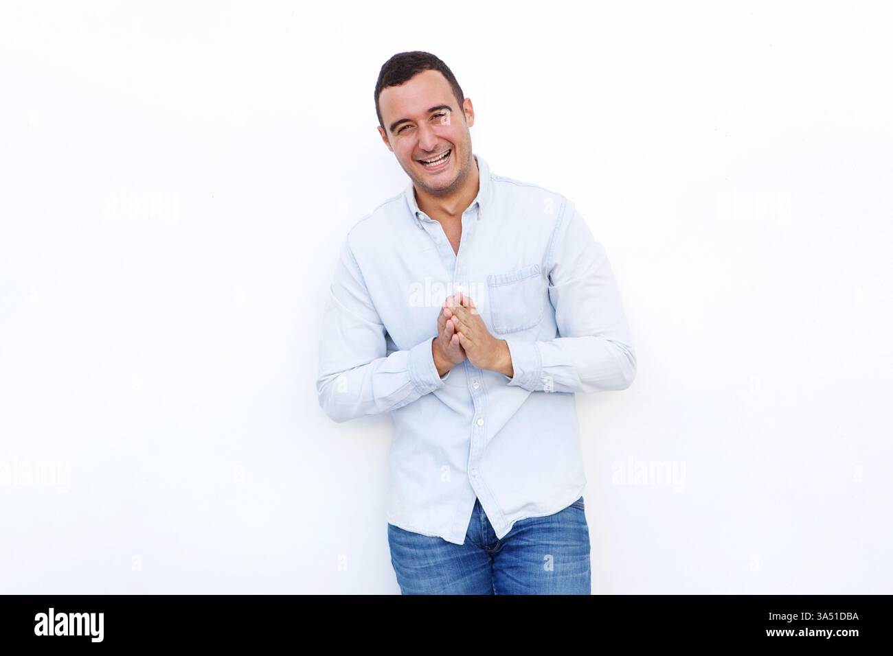 Joyeux homme hispanique se tient contre un fond blanc propre de studio avec ses mains serrées, projetant la confiance et l'accessibilité. Parfait pour les campagnes lifestyle et mode mettant en vedette un homme latino décontracté et moderne. Idéal pour les publicités et les sites Web présentant des images de personnages optimistes. Banque D'Images