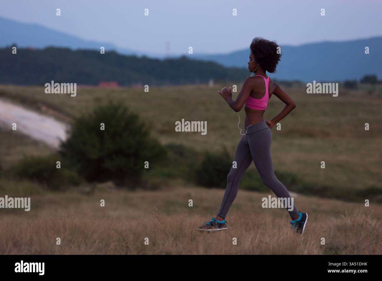 Jeune coureuse noire avec écouteurs jogging en plein air dans la nature lors d'une belle nuit d'été. Cette image de fitness met en évidence l'activité extérieure et une vie saine. Convient pour les campagnes sur la course à pied, l'entraînement et le bien-être estival. Banque D'Images