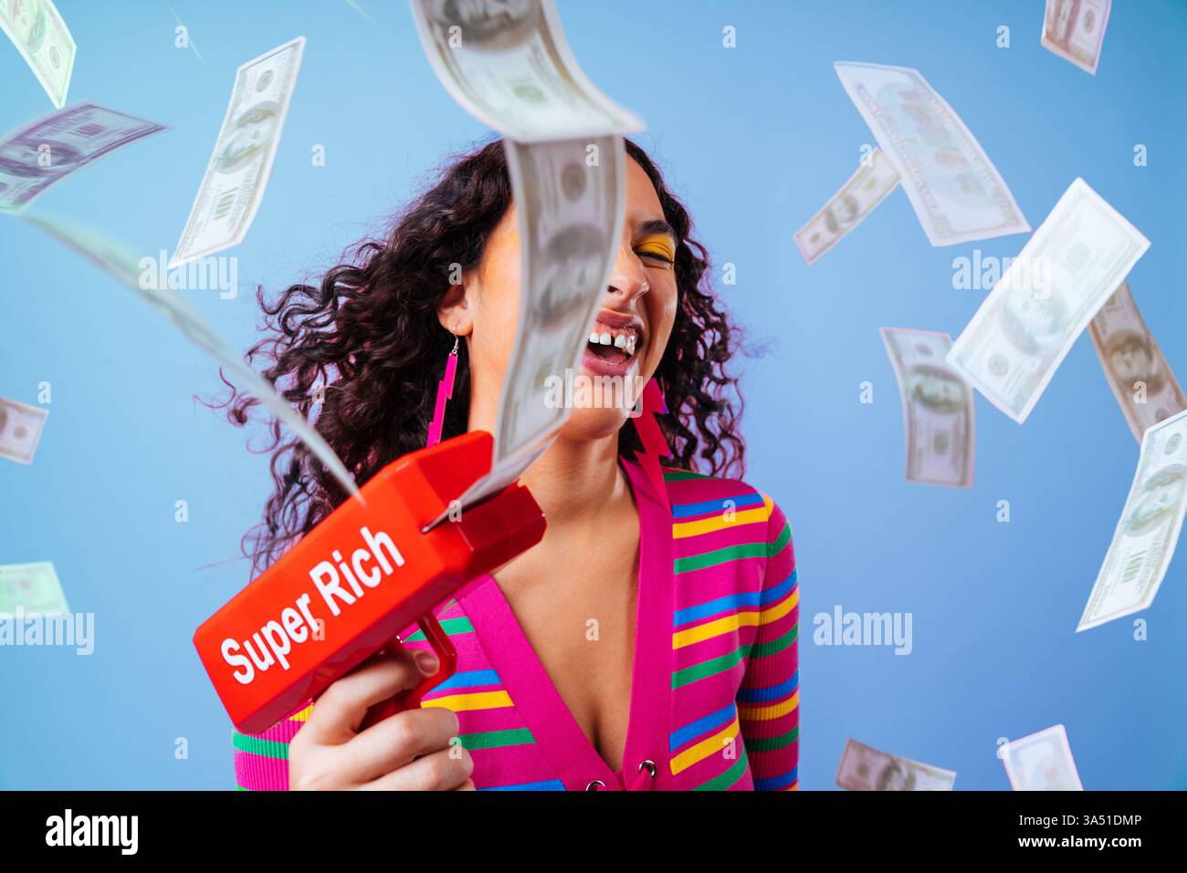 Joyeuse femme hispanique avec diastema tenant l'arme à argent debout avec des billets d'un dollar volants sur fond bleu Banque D'Images