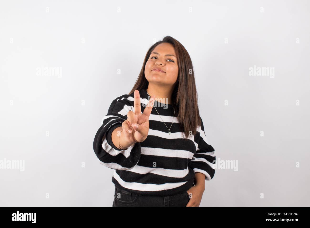 Portrait d'une femme hispanique joyeuse montrant un signe de paix sur un fond de studio gris. Une image élégante et optimiste idéale pour les thèmes de style de vie, de mode et d'autonomisation. Banque D'Images