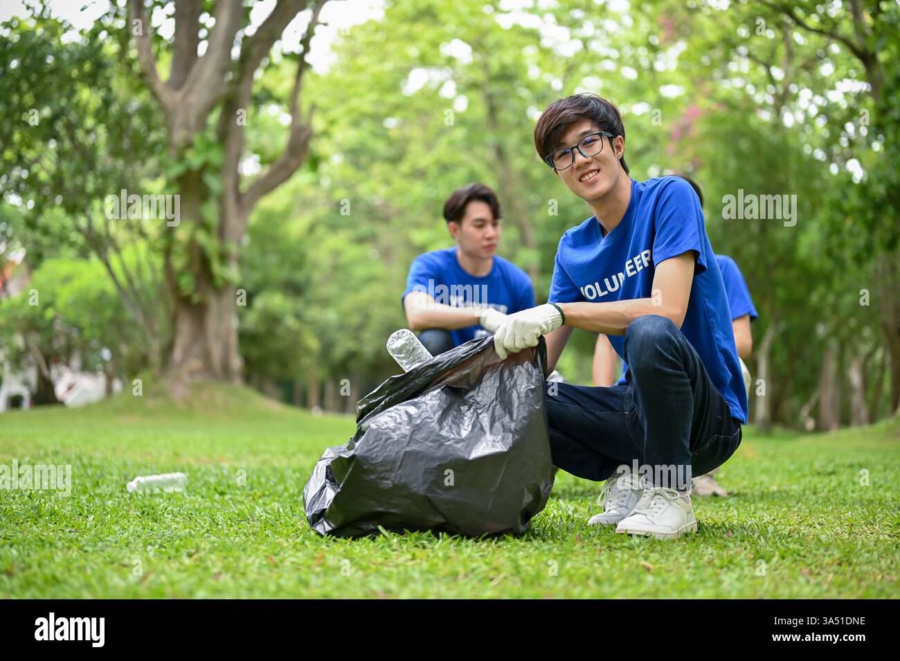 Un jeune volontaire asiatique heureux avec un sac poubelle en plastique nettoie un parc public avec son équipe. L'image met en valeur le service communautaire et la protection de l'environnement. Idéal pour le bénévolat, la RSE et les campagnes de sensibilisation à l'environnement. Banque D'Images