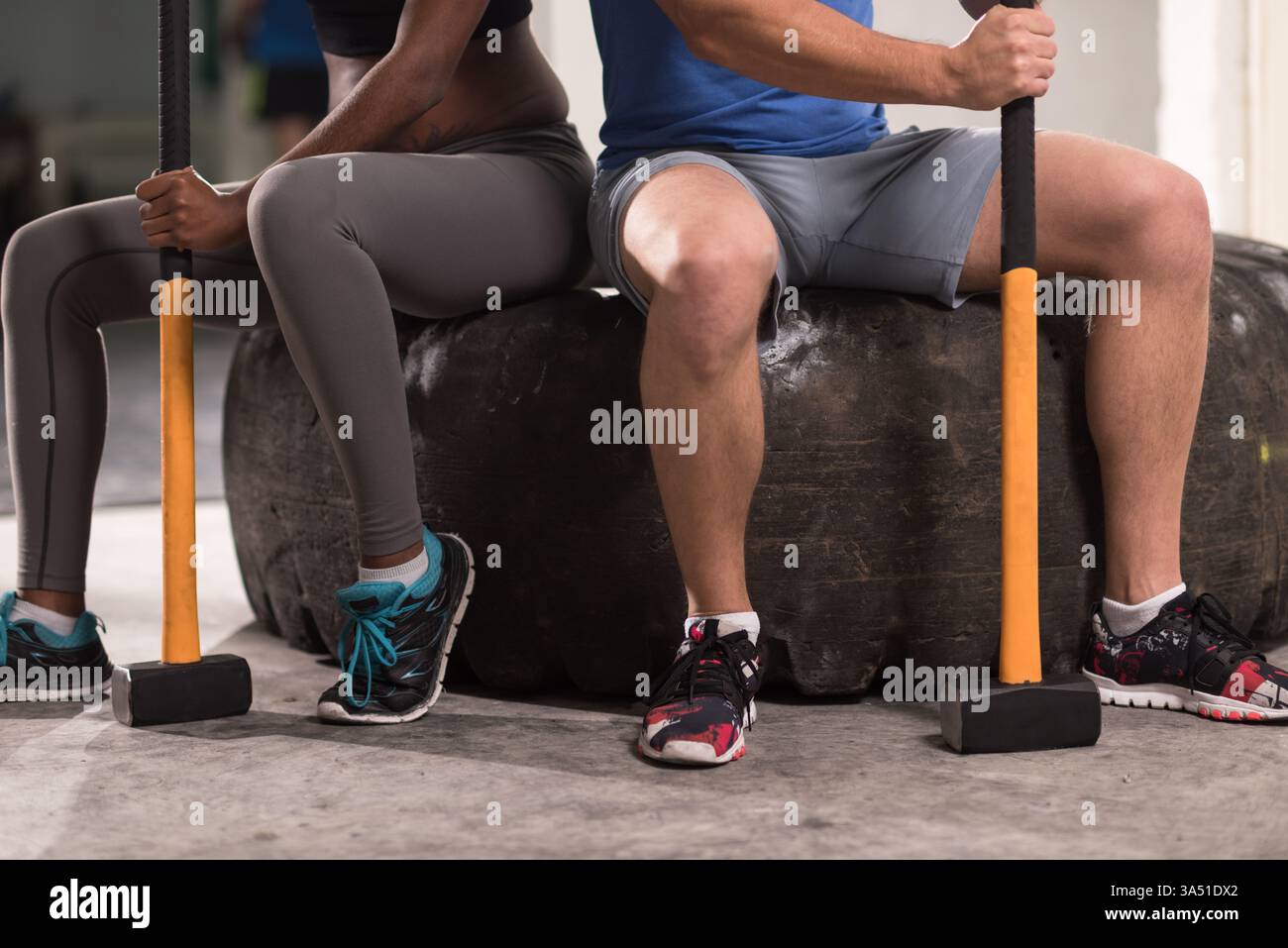 Monter couple dans l'usure athlétique tenir des marteaux de traîneau tout en étant assis sur un pneu de tracteur dans un cadre de gymnastique. Cette image dynamique exprime la motivation de l'entraînement en force et de l'entraînement pour les campagnes de fitness. Banque D'Images