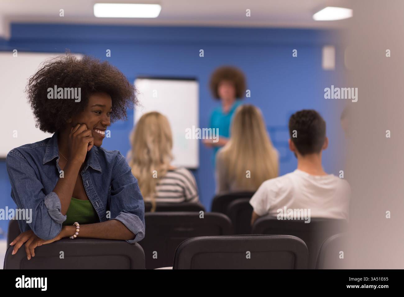 Femme noire joyeuse avec un afro reposant son visage dans sa main, assise en arrière sur une chaise à côté d'un groupe de gens d'affaires dans une salle de conférence. Adapté aux thèmes du lieu de travail, de la diversité et du travail d'équipe dans les communications d'entreprise. Idéal pour les réunions, les bureaux et les contextes de style de vie professionnel. Banque D'Images
