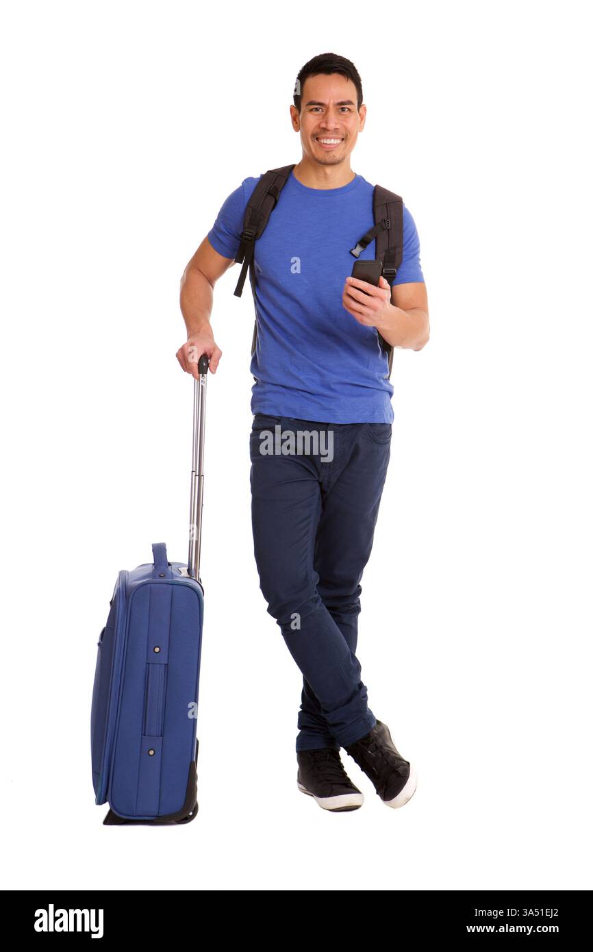 Homme asiatique souriant avec des bagages tenant smartphone debout avec des jambes croisées sur fond blanc Banque D'Images