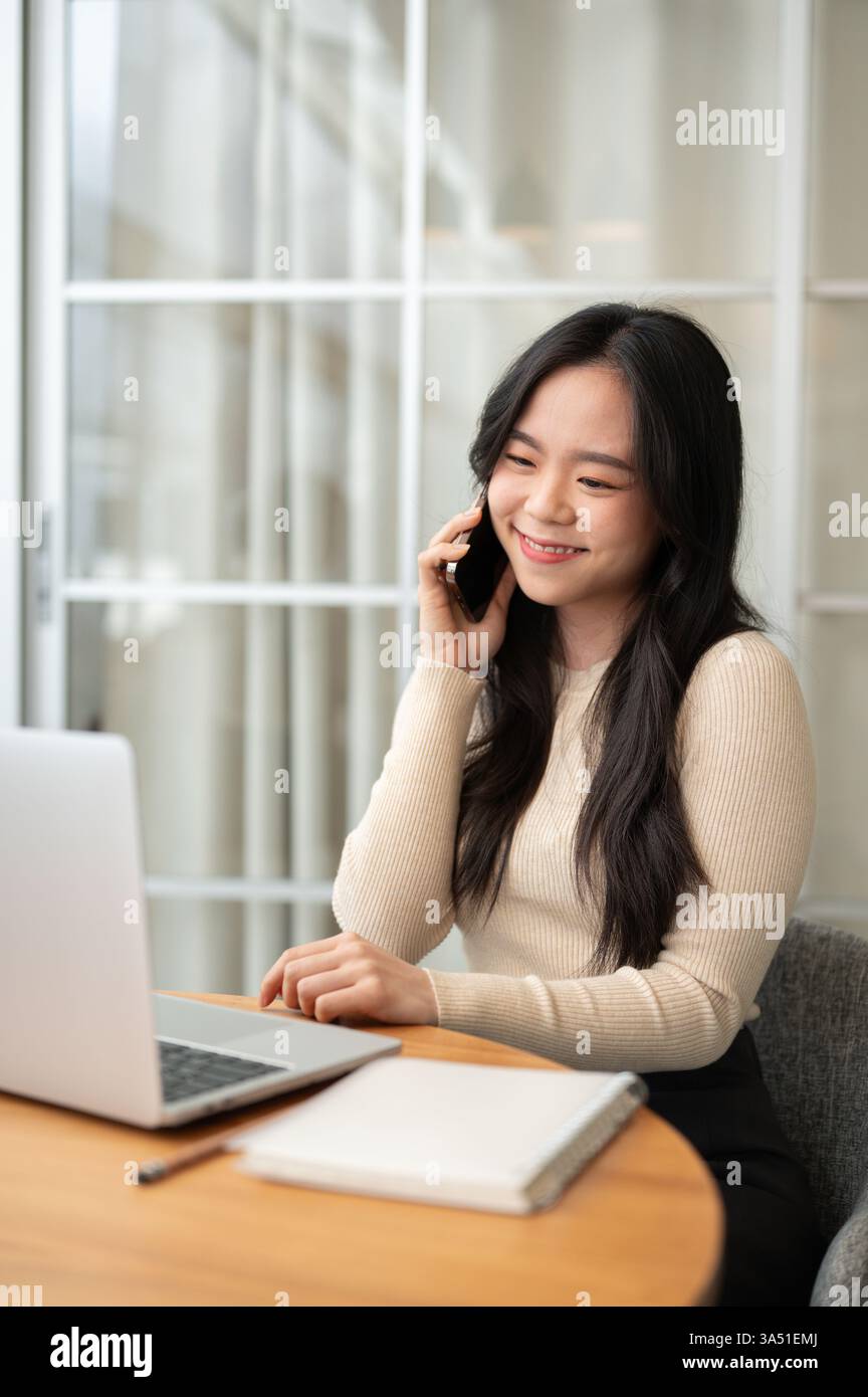 Femme asiatique souriante regardant un ordinateur portable tout en ayant un appel téléphonique assis à table à la maison Banque D'Images