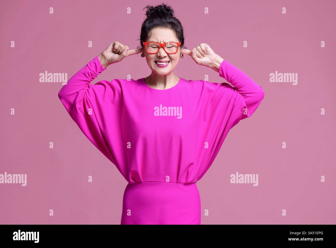 Femme chinoise senior couvre ses oreilles pour bloquer le bruit dans un portrait de studio avec un fond rose. L'image transmet l'irritation des sons forts et convient aux sujets de bruit, de concentration et de stress. À utiliser pour le contenu éditorial ou lifestyle sur la sensibilité sonore. Banque D'Images