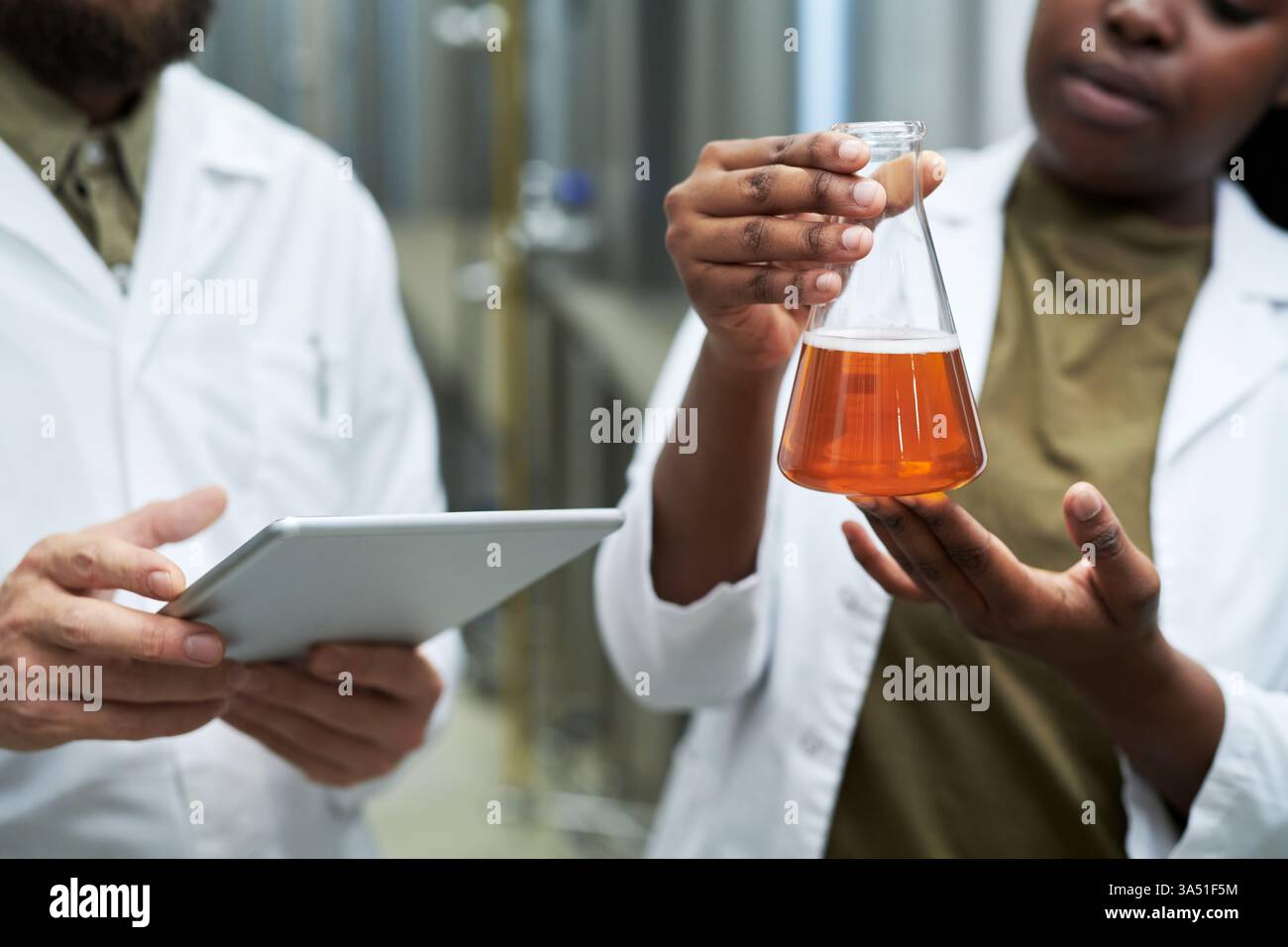 Une équipe diversifiée de techniciens de laboratoire vérifie la qualité de la bière dans une microbrasserie. Portant des blouses de laboratoire, ils utilisent de l’équipement et une tablette numérique pour surveiller la fermentation et assurer une production de bière homogène. Banque D'Images