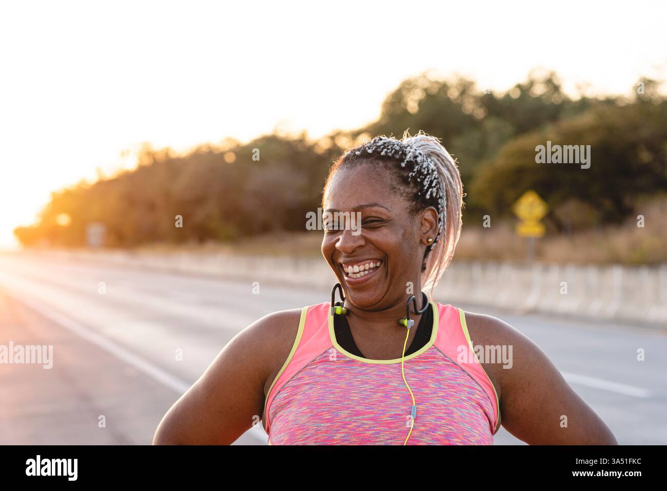 Femme afro-latina joyeuse en vêtements actifs riant avec les mains sur les hanches au coucher du soleil sur le bord de la route. Idéal pour le fitness, les activités de plein air et les campagnes de vie saine. Utilisation dans le bien-être ou l'image de marque sportive pour la motivation et la vitalité. Banque D'Images