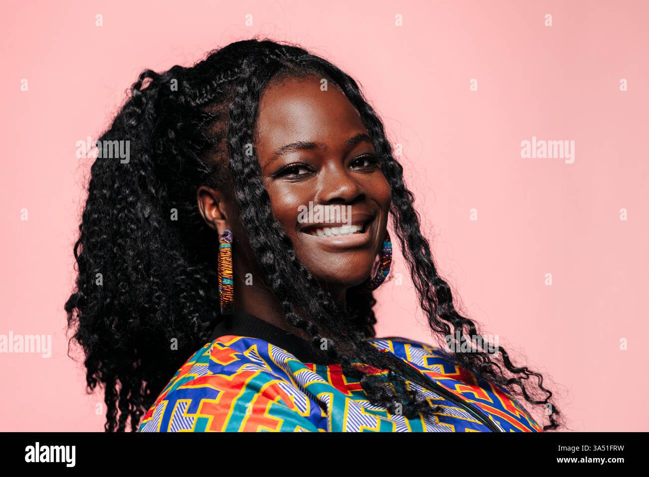 Femme afro-américaine souriante avec les cheveux bouclés se tient contre un mur rose. Image de style de vie adaptée aux portraits, parfaite pour la mode, la beauté et les thèmes féminins confiants. Idéal pour les soins personnels, le marketing boutique et les campagnes de médias sociaux. Banque D'Images