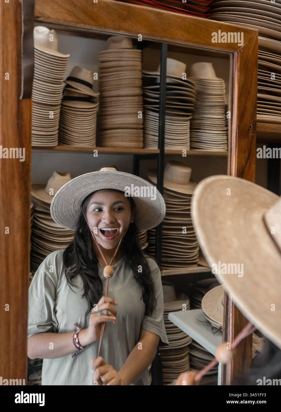 Jeune femme hispanique regarde dans le miroir tout en essayant un nouveau chapeau dans une boutique. La scène capture le shopping de mode, le style latin, et l'exploration de style personnel. Parfait pour la vente au détail de mode, style de vie et contenu culturel. Banque D'Images