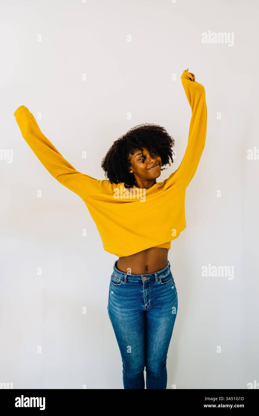 Femme hispanique souriante en denim et un pull jaune se tient à l'intérieur près d'un mur blanc avec les bras tendus. Ses cheveux bouclés et son style décontracté donnent un portrait gai de son style de vie. Banque D'Images