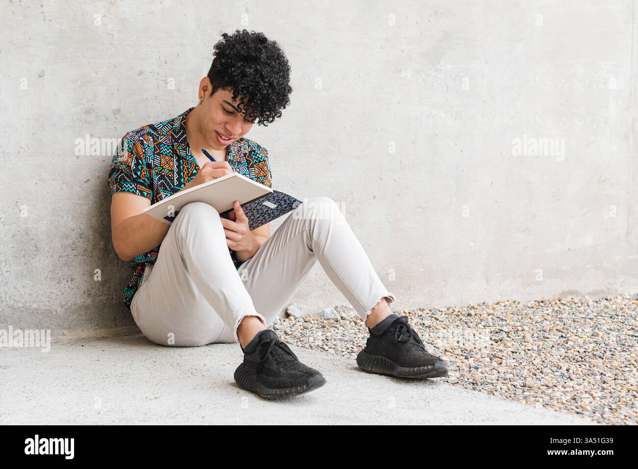 Portrait intégral d'un vénézuélien vêtu de vêtements décontractés, assis sur un sol en béton à l'extérieur tout en écrivant dans un cahier. Cette scène est parfaite pour le contenu sur la journalisation, l'étude et la créativité personnelle dans les environnements quotidiens. Banque D'Images