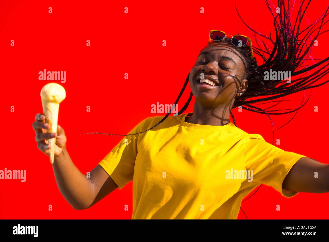 Image vibrante d'une jeune femme africaine élégante dansant et dégustant de la crème glacée sur un fond rouge audacieux. Idéal pour les campagnes estivales de style de vie, de mode et de nourriture et de boissons. La pose énergique communique le bonheur, l'espiègle et le plaisir insouciant. Banque D'Images