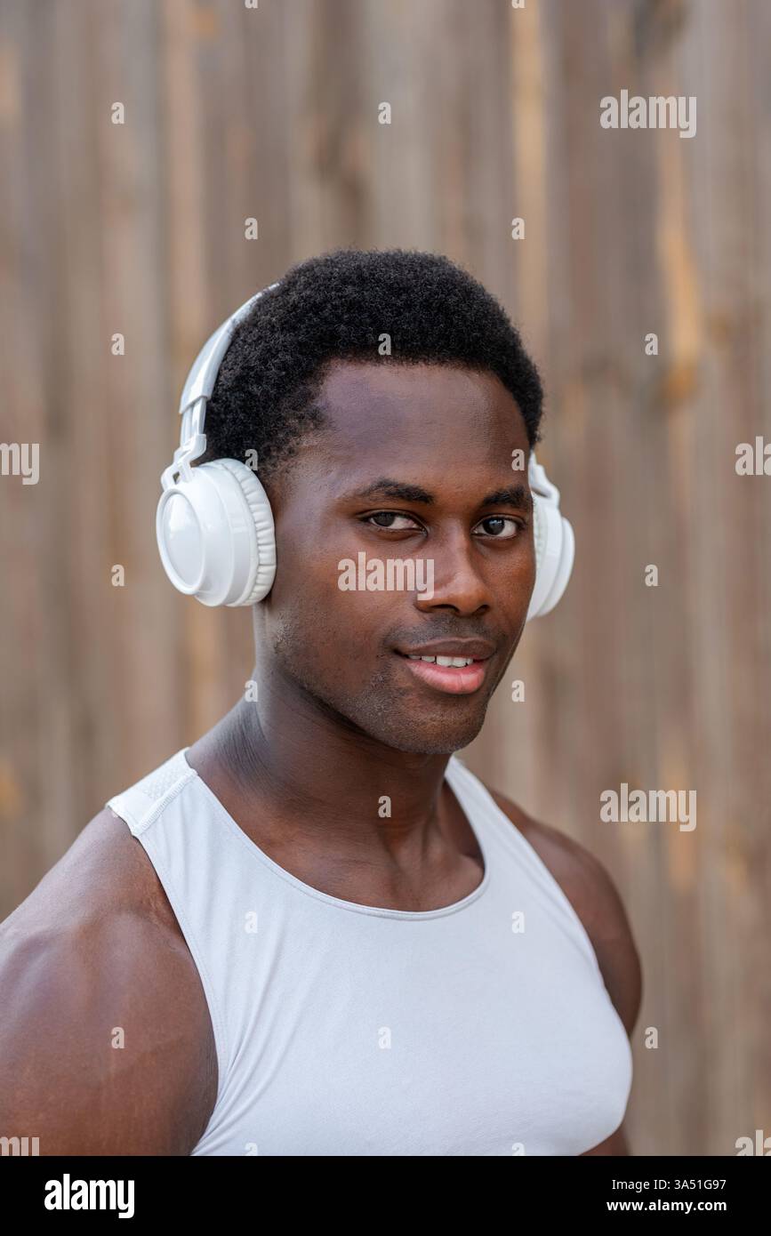 Portrait d'un jeune athlète masculin noir musclé portant un haut blanc et des écouteurs sans fil pendant un entraînement en plein air de jour. Il regarde la caméra avec confiance, transmettant énergie, mise au point et détermination. Idéal pour les campagnes de fitness, les vêtements de sport et la marque de bien-être avec une ambiance athlétique. Banque D'Images