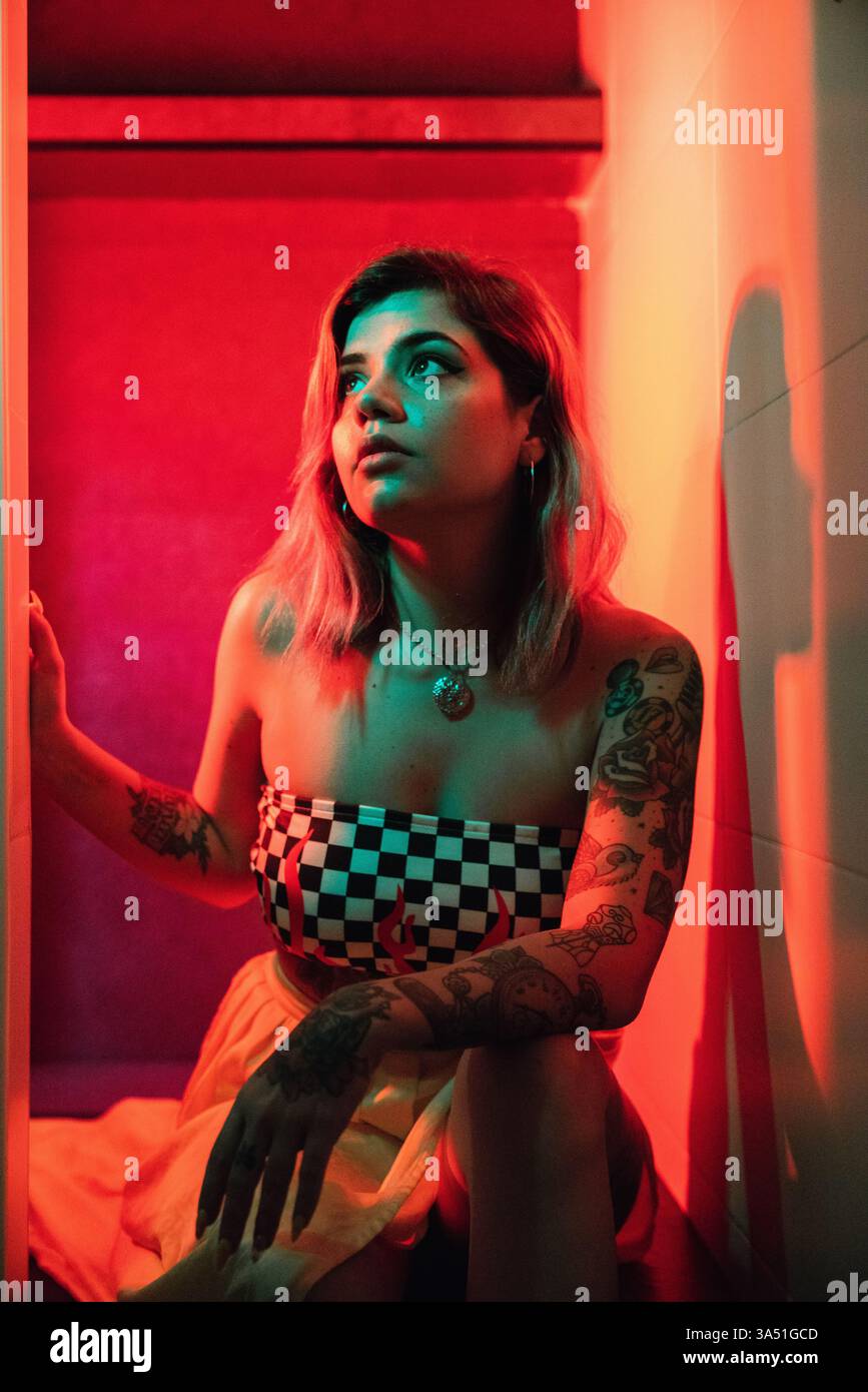 Une femme hispanique tatouée est assise sur le sol dans une pièce lumineuse et éclairée, regardant loin avec son bras reposant sur son genou. Cette image de style de vie met en valeur un moment de détente, à la maison, idéal pour la vie à la maison, le style personnel et les thèmes de travail créatifs. Parfait pour les campagnes autour de l'individualité, de l'expression de soi et de la vie moderne. Banque D'Images