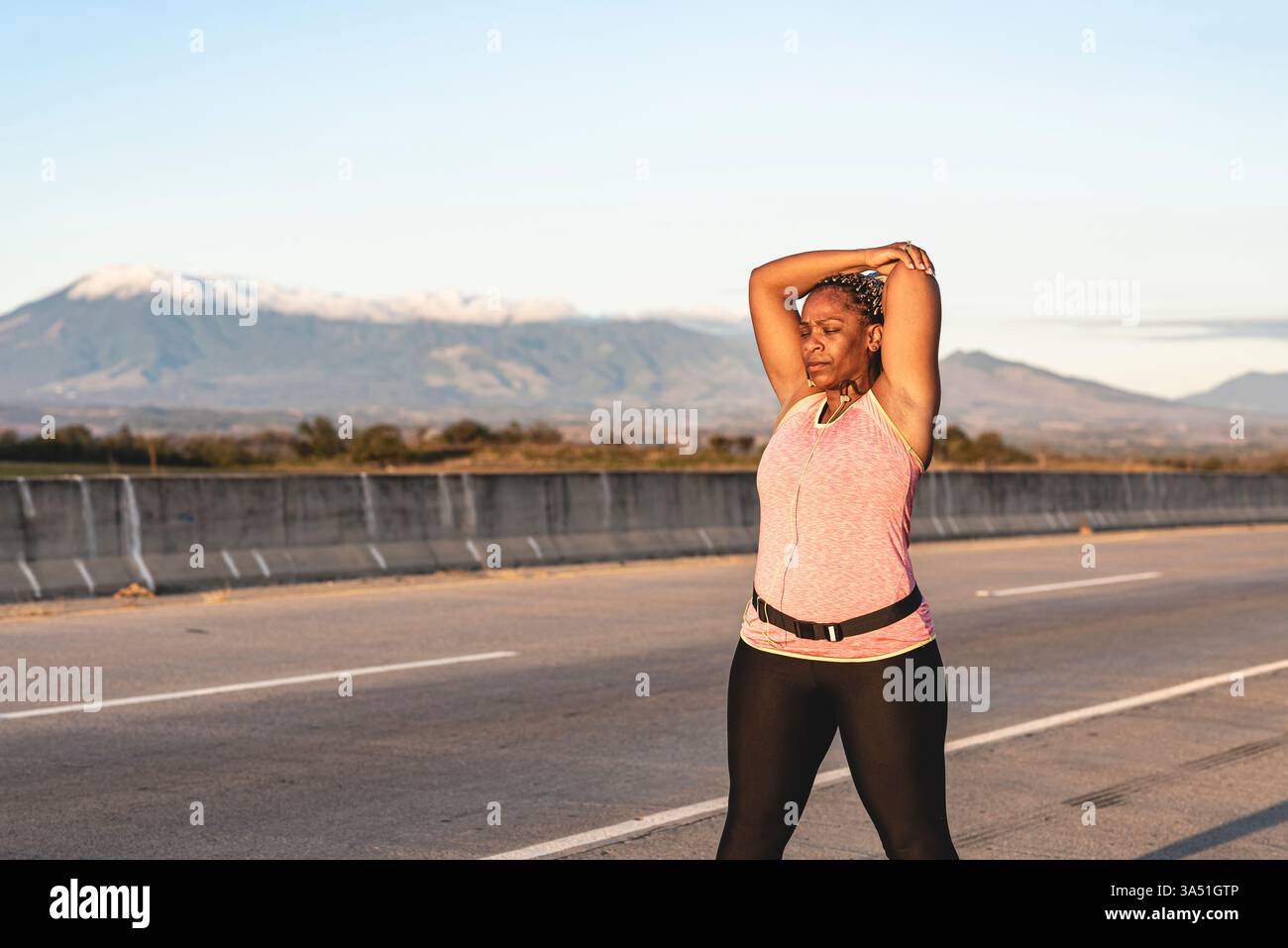 Femme afro-latina en vêtements actifs s'étire avec les yeux fermés sur le bord de la route au coucher du soleil. Le moment de remise en forme en plein air met l'accent sur la santé, la vitalité et la concentration, adapté aux campagnes de bien-être et de sport. Idéal pour des images inclusives sur un style de vie actif mettant en scène des publics variés. Banque D'Images