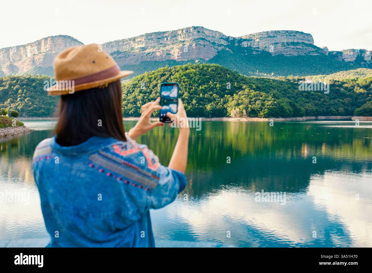Jeune femme latina debout sur un rebord au bord du lac, prenant une photo avec un smartphone. Une scène de style de vie en plein air lumineuse parfaite pour les voyages, la photographie ou les campagnes sur les réseaux sociaux. Idéal pour le tourisme, les marques de plein air ou la marque personnelle. Banque D'Images
