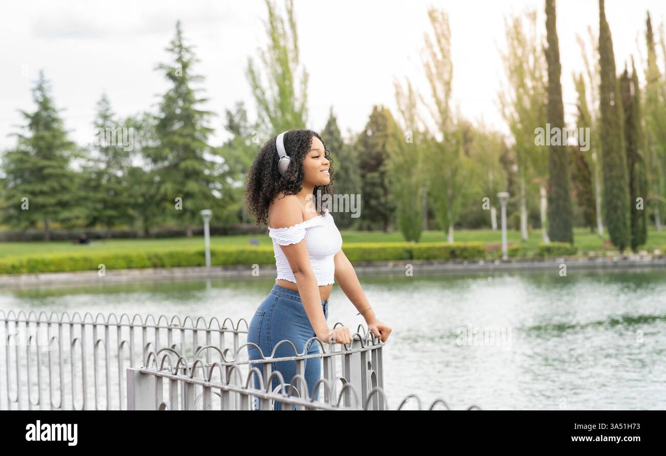 Femme noire portant des écouteurs se penche sur une clôture dans un parc ensoleillé. Le moment de détente et de style de vie convient à la musique, aux loisirs en plein air et aux thèmes du bien-être. Parfait pour les campagnes sur la nature urbaine, le plaisir personnel et la vie contemporaine. Banque D'Images