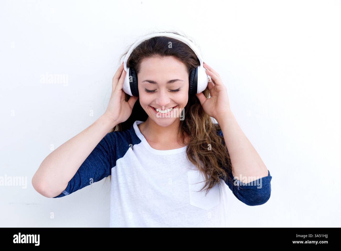 Femme asiatique souriante avec des écouteurs se tient sur un fond blanc, écoutant de la musique. Ambiance décontractée et insouciante parfaite pour les sujets de style de vie et de divertissement. Idéal pour les fonctionnalités sur la musique, la détente et le style personnel moderne. Banque D'Images