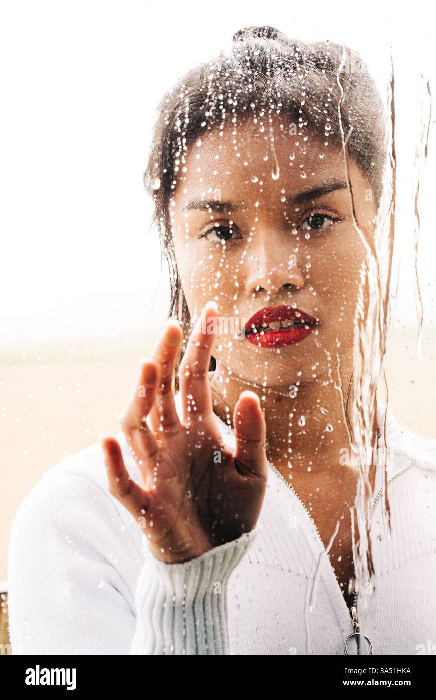 Une femme asiatique se tient derrière un verre avec des gouttes de pluie à la surface dans un studio. Son humeur réfléchie et le réglage de la fenêtre pluvieuse de humeur sombre rendent ce portrait adapté à la beauté, à la mode ou aux traits de style de vie. L'éclairage lumineux du studio contraste avec l'humidité sur la fenêtre pour créer une image dramatique. Banque D'Images