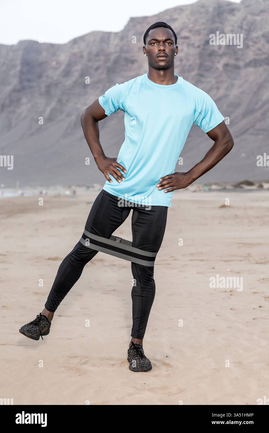 Homme afro-américain effectue un exercice de jambe sur la plage, les mains sur ses hanches en tenue athlétique. Ce cliché de fitness en plein air transmet la vitalité et un style de vie actif au bord de la mer. Banque D'Images