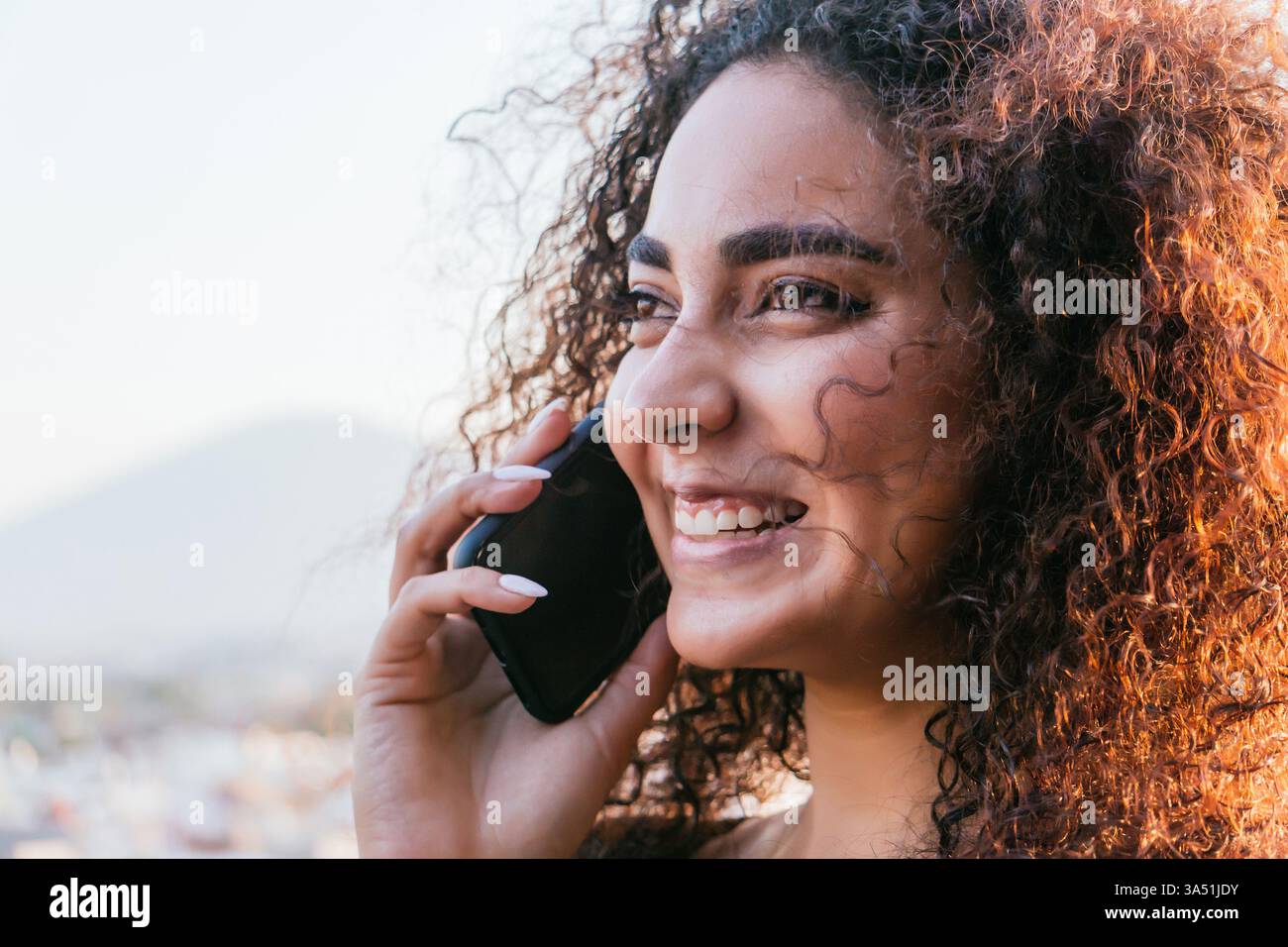 Femme hispanique souriante utilisant un smartphone tout en se tenant dans une rue urbaine au coucher du soleil. Cette scène de rue dynamique est idéale pour les sujets sur la communication mobile, la vie sociale et l'utilisation quotidienne de la technologie. La lumière chaude et le sourire joyeux créent une ambiance positive et accessible. Banque D'Images