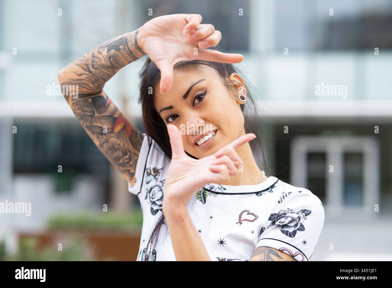 Femme asiatique avec un bras tatoué sourit tout en encadrant un geste de tir. L'image transmet une ambiance urbaine moderne et un style personnel. Idéal pour les portraits, la mode et le contenu de style de vie. Banque D'Images