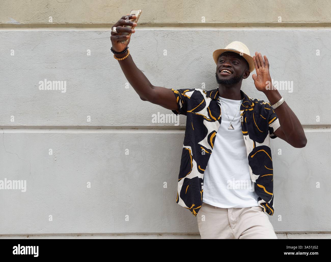 Élégant jeune homme afro-américain dans une chemise colorée et chapeau vagues et prend un selfie près d'un mur de pierre dans la ville. Image décontractée et optimiste, idéale pour les campagnes sur les réseaux sociaux, la technologie mobile et le style de vie urbain. Idéal pour le contenu sur le style personnel, les selfies et la mode de rue. Banque D'Images