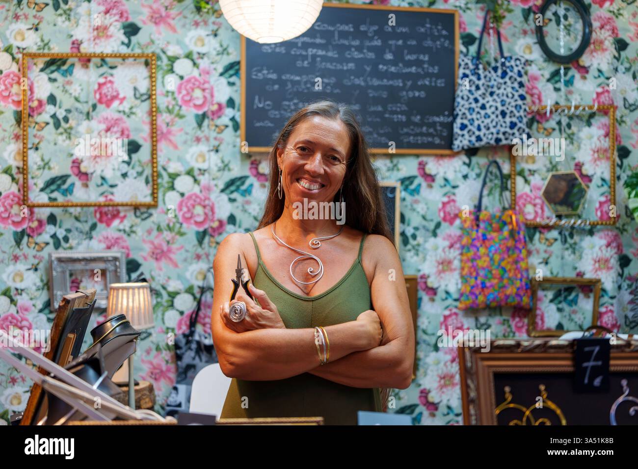 Fière artisan hispanique se tient à son stand de rue avec des outils à la main, vendant des bijoux en fil d'aluminium faits à la main. Une scène de marché en plein air qui célèbre les petites entreprises, l'artisanat et l'esprit d'entreprise. Banque D'Images