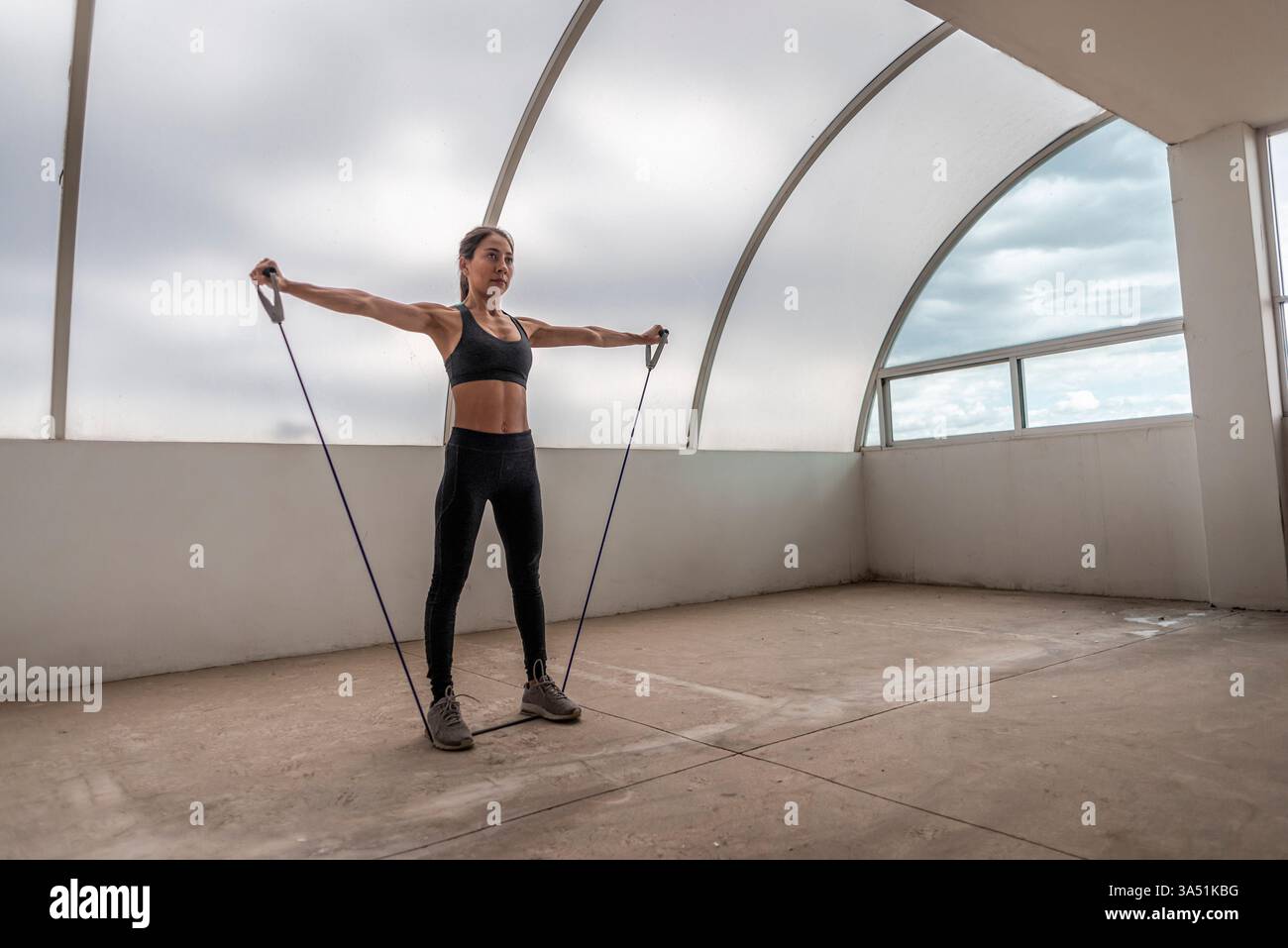 Femme hispanique en vêtements de sport effectuant une séance d'entraînement avec une bande de résistance sur un toit pendant la journée. Parfait pour le fitness, l'entraînement en plein air et les campagnes de vie saine. La scène lumineuse et énergique convient à la marque sportswear et lifestyle. Banque D'Images