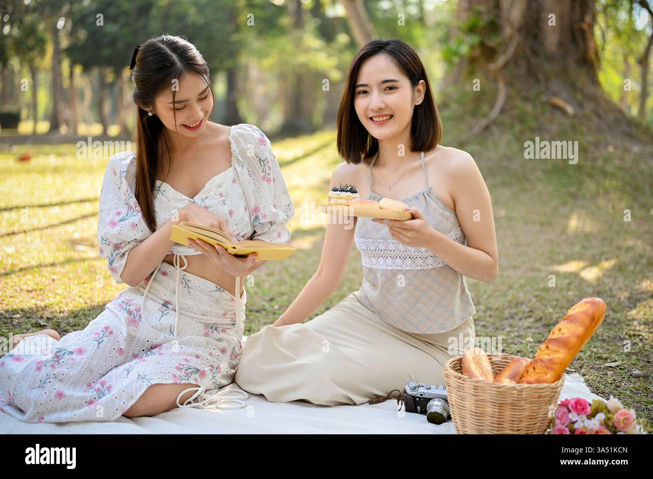 Couple féminin asiatique souriant lisant un livre et partageant un beignet sur une couverture de parc pendant la journée. Ce moment convivial LGBTQ+ capte l’amitié, la détente et les loisirs en plein air. Idéal pour les campagnes autour de l'amour, de la communauté et des moments quotidiens entre amis. Banque D'Images