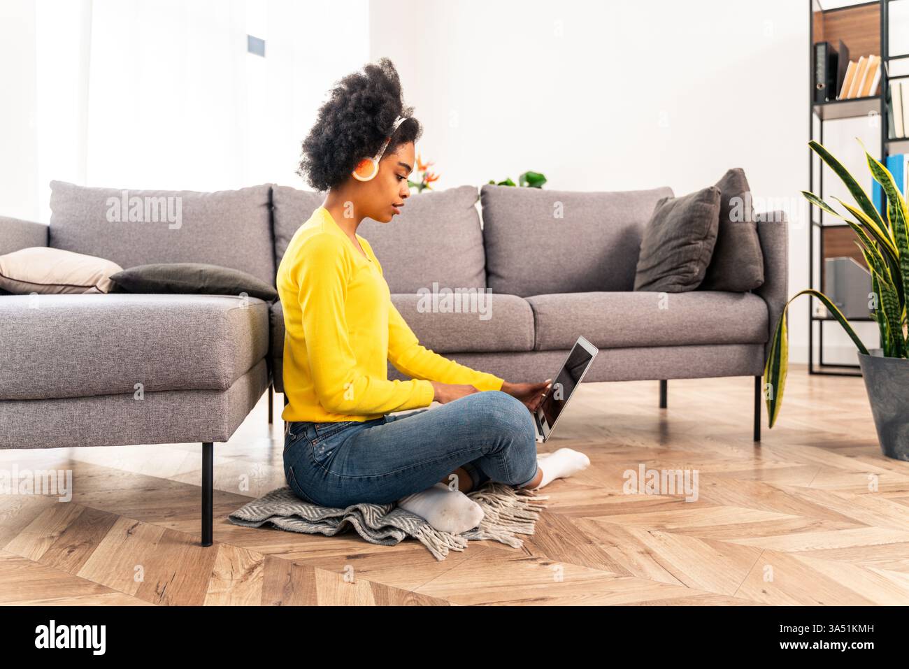 Portrait d'une femme afro-américaine élégante à la maison, portant des écouteurs et utilisant un ordinateur portable pour la musique en ligne et le travail à distance. La configuration décontractée du salon avec un ordinateur portable moderne et des écouteurs donne une ambiance contemporaine de bureau à domicile et de divertissement. Idéal pour les campagnes sur le travail à distance, l'étude à domicile, l'apprentissage en ligne ou la musique en streaming. Banque D'Images