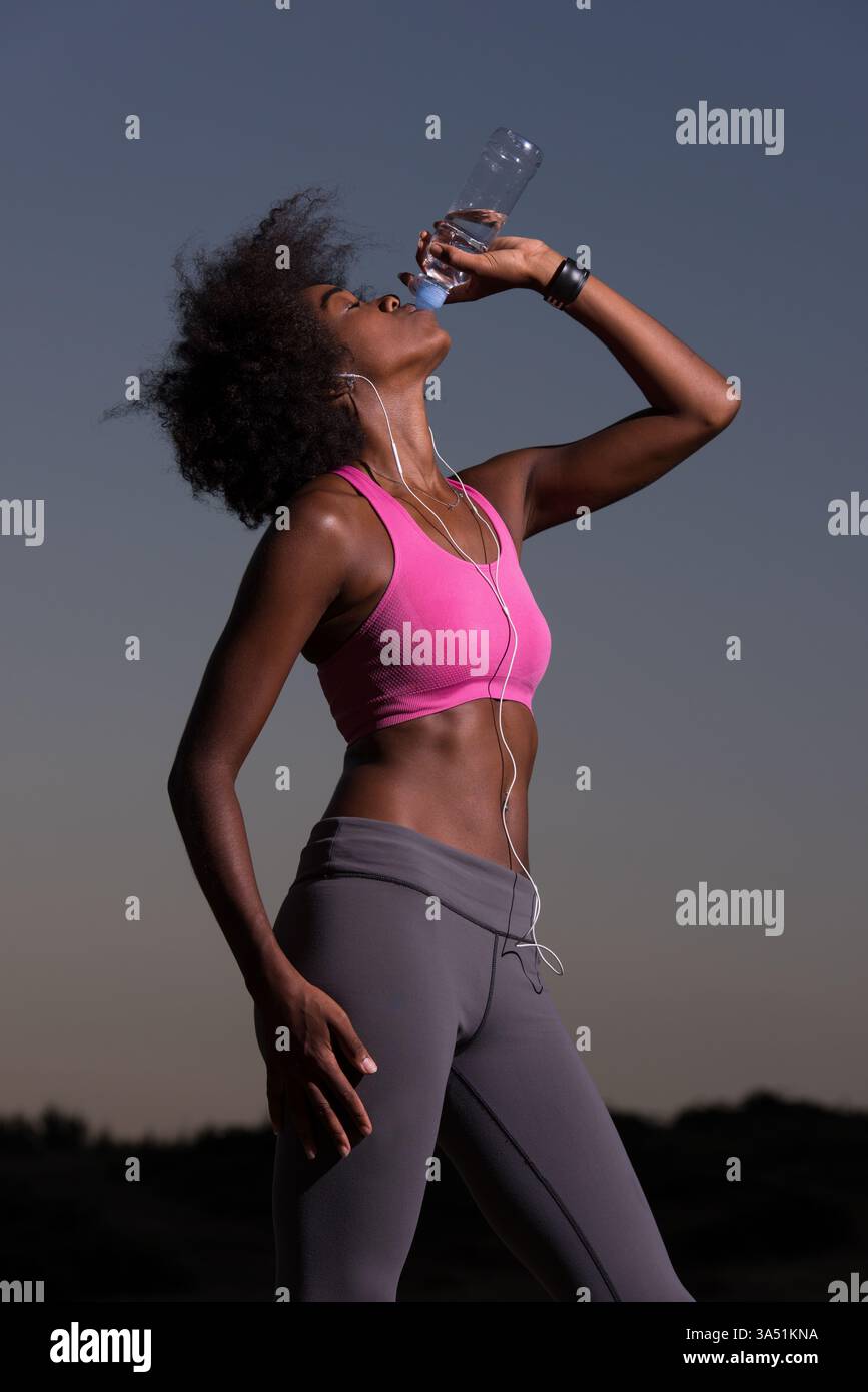 Jeune femme afro-américaine avec des écouteurs boit de l'eau après avoir fait du jogging dans un pré au crépuscule. Elle incarne un mode de vie sain et actif dans un cadre de nuit d'été. Parfait pour les campagnes de fitness, de bien-être et d'aventure en plein air. Banque D'Images