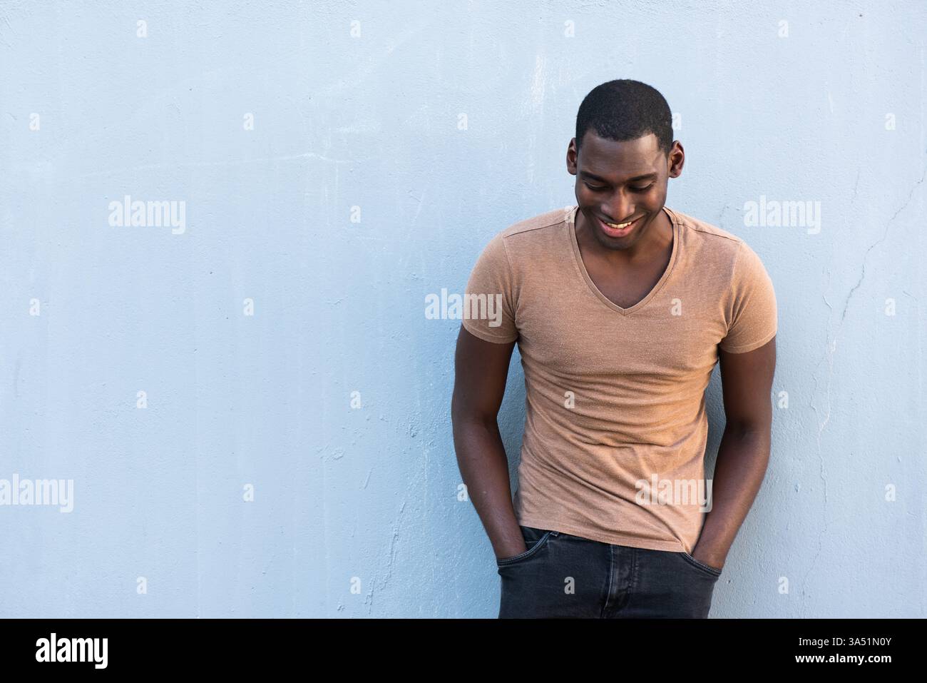 Homme noir souriant avec les mains dans les poches se tient sur un fond gris, regardant vers le bas. Un portrait polyvalent adapté aux campagnes de style de vie, de mode et de style personnel. Banque D'Images