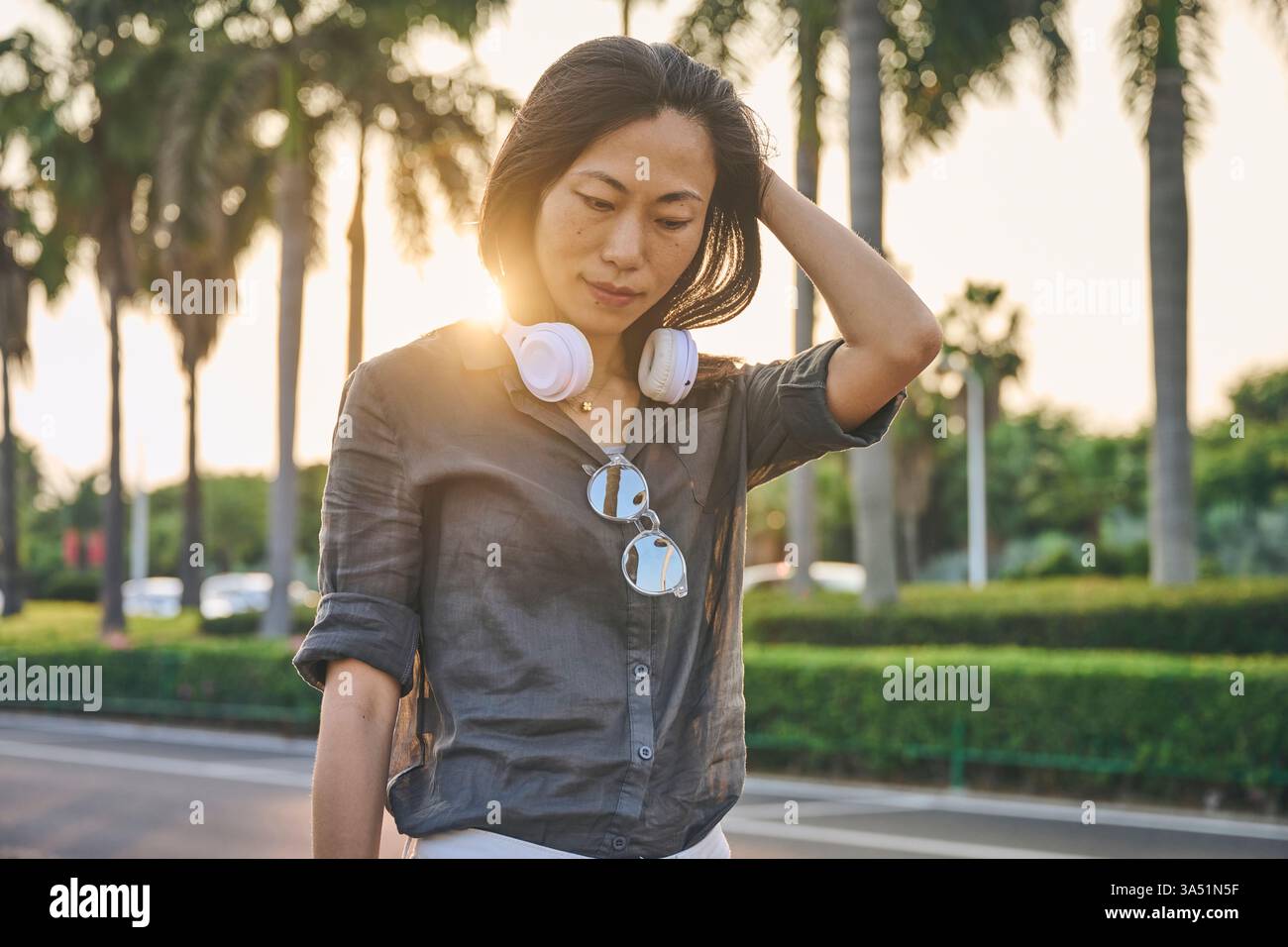Femme asiatique dans une rue ensoleillée portant des écouteurs et passant une main dans ses cheveux. Un portrait élégant de style de vie qui convient à la musique, à la mode et aux thèmes de la vie urbaine. Idéal pour les campagnes visant la culture numérique et le style personnel. Banque D'Images