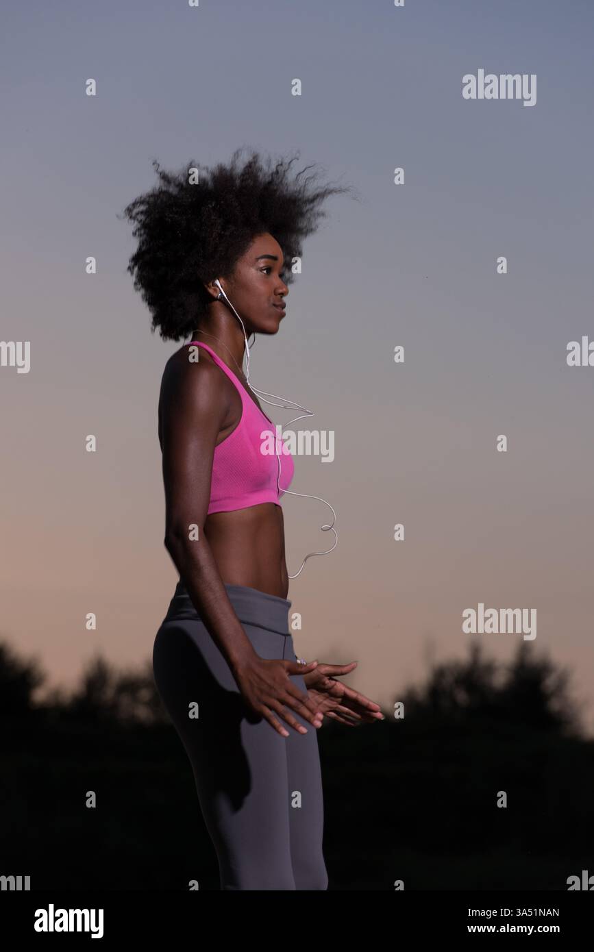 Jeune femme noire en bonne santé effectue un exercice d'étirement après un jogging dans une prairie lors d'une belle soirée d'été. Le moment de détente et d'athlétisme correspond à des thèmes de fitness, de course à pied et de style de vie en plein air. Banque D'Images