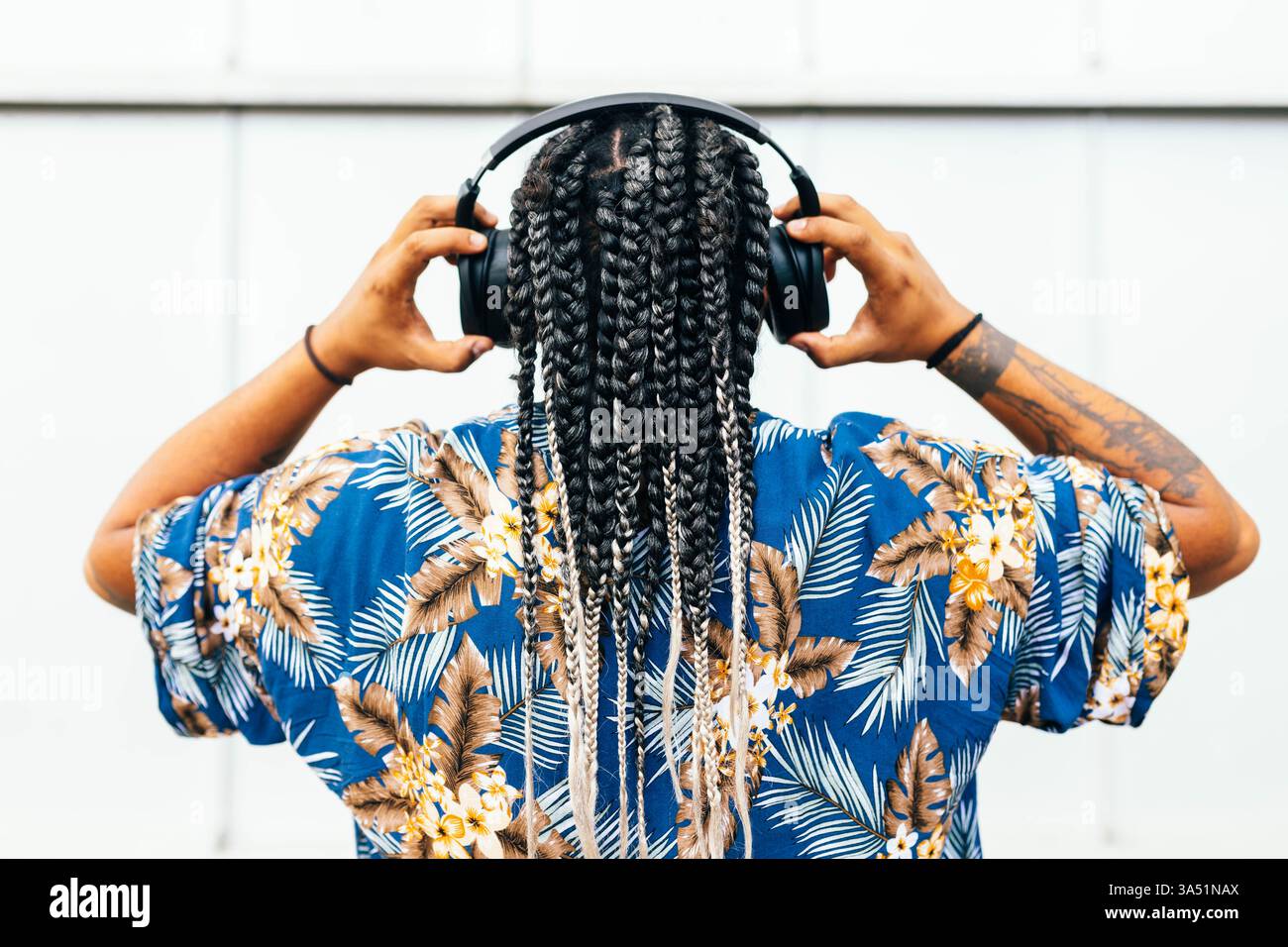 Homme latin vu de derrière, portant une chemise bleue florale avec des tresses et des tatouages visibles, écoutant de la musique sur des écouteurs noirs. Un look contemporain idéal pour la musique, les écouteurs et les campagnes de Street fashion. La perspective arrière ajoute une ambiance urbaine et détendue avec une attention particulière au style personnel. Banque D'Images