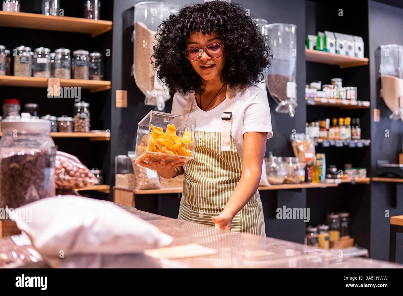 Bonne jeune femme afro-américaine en tablier rayé et des verres sourire et mettre boîte de verre de mangue séchée sur l'étagère pendant le travail dans zéro gaspillage s épicerie Banque D'Images
