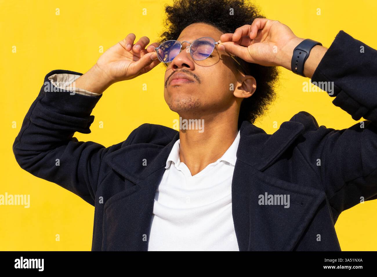 Homme hispanique avec les yeux fermés touchant ses lunettes contre un mur jaune vif. Un portrait élégant qui met l'accent sur les lunettes, l'individualité et la mode contemporaine. Idéal pour le style de vie, la mode et les thèmes de marque personnelle confiants. Banque D'Images