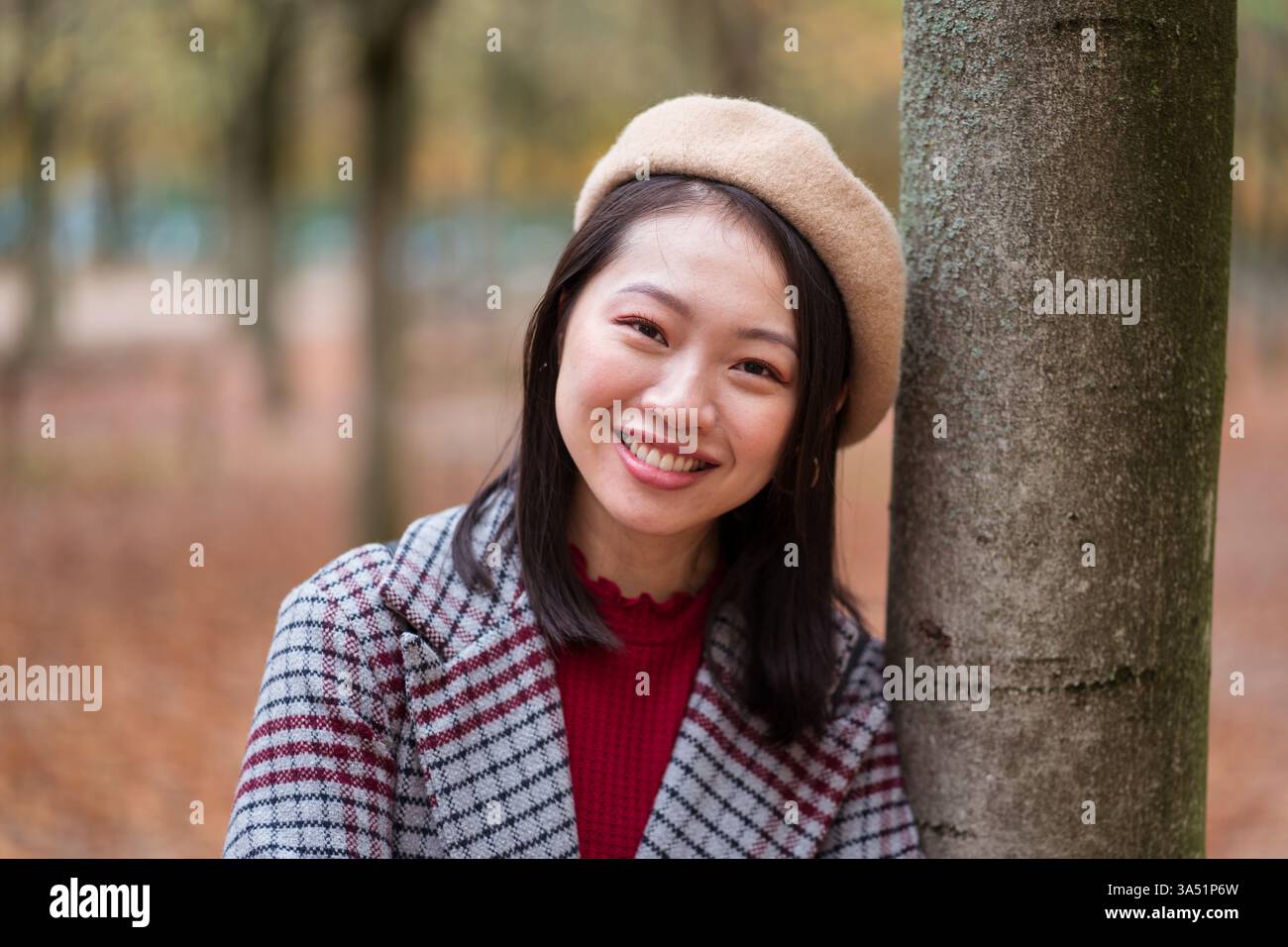 Femme asiatique souriante dans un béret et un manteau à carreaux s'appuie sur un arbre dans un parc d'automne. Ce portrait saisonnier fonctionne bien pour la mode, le style de vie et le marketing saisonnier. Idéal pour une utilisation éditoriale ou commerciale avec un look extérieur élégant et confiant. Banque D'Images