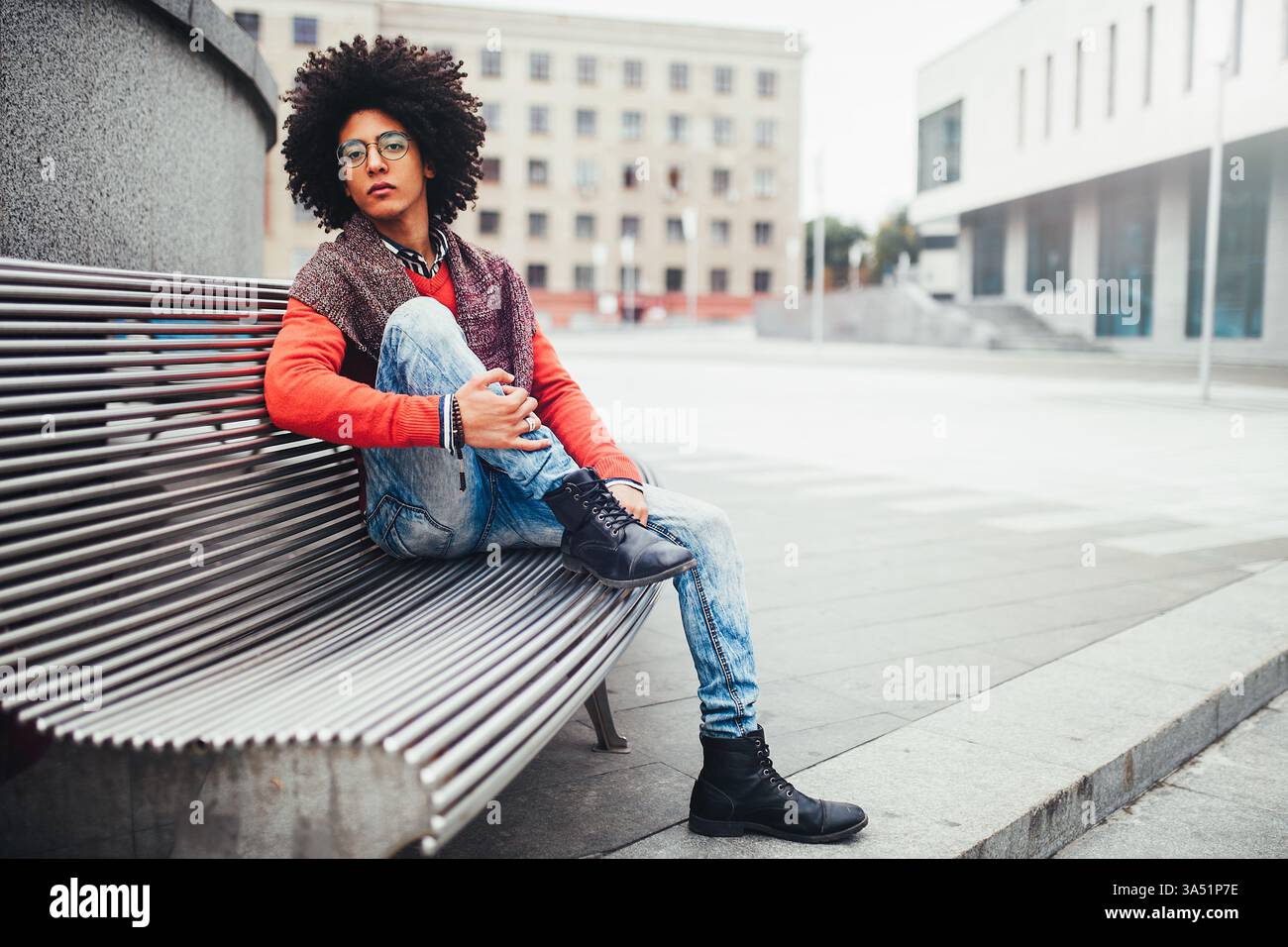 Un bel étudiant égyptien aux cheveux bouclés est assis sur un banc dans un cadre urbain, portant un pull orange vif et un Jean. Le moment capture un style extérieur décontracté et des pauses d'étude entre les leçons. Banque D'Images
