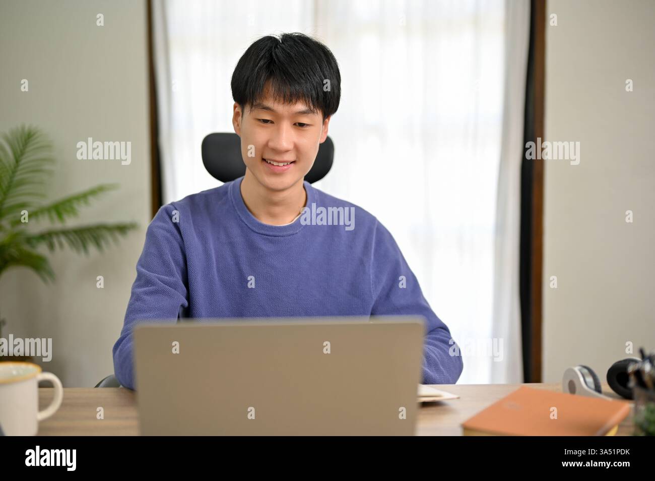 Jeune étudiant asiatique dans un pull confortable utilise un ordinateur portable à la maison, axé sur un projet scolaire ou une classe en ligne. Idéal pour les thèmes de l'étude à distance, de l'apprentissage en ligne et de la vie étudiante. Banque D'Images