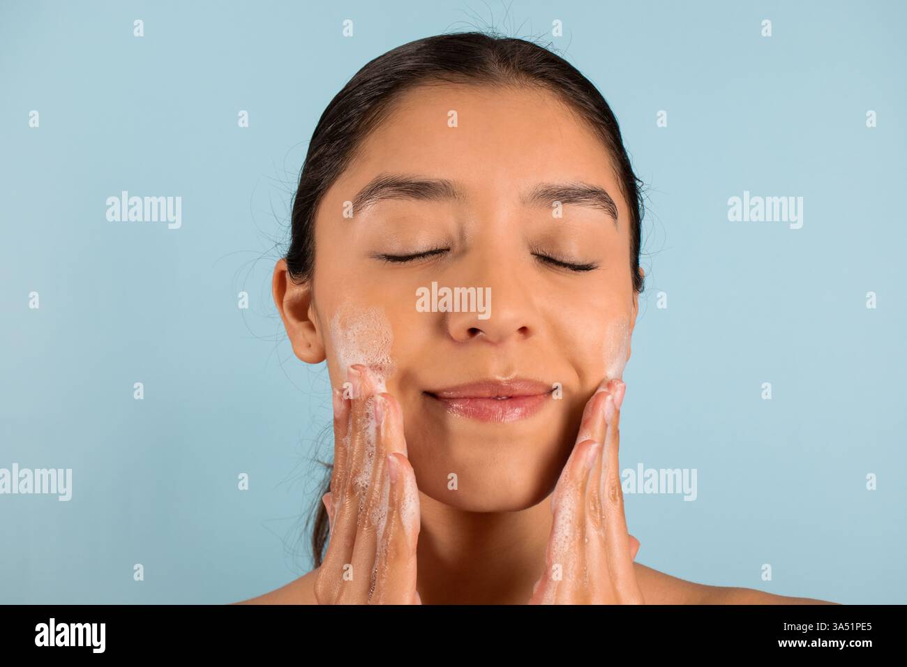 Vue de face d'une femme hispanique les yeux fermés, appliquant un produit de soin mousseux sur son visage sur un fond bleu. Ce moment de beauté communique les soins personnels, le bien-être et les soins de routine de la peau. Convient pour les cosmétiques, les marques de soins de la peau et le contenu dermatologique. Banque D'Images