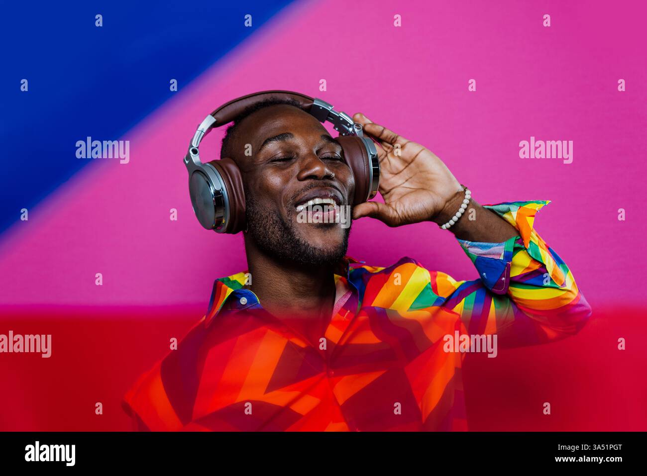 Homme afro-américain souriant chantant tout en écoutant de la musique, portant des écouteurs, avec des lumières de gel colorées en arrière-plan. Un portrait studio dynamique idéal pour la musique, la performance et les campagnes de style de vie créatif. Banque D'Images