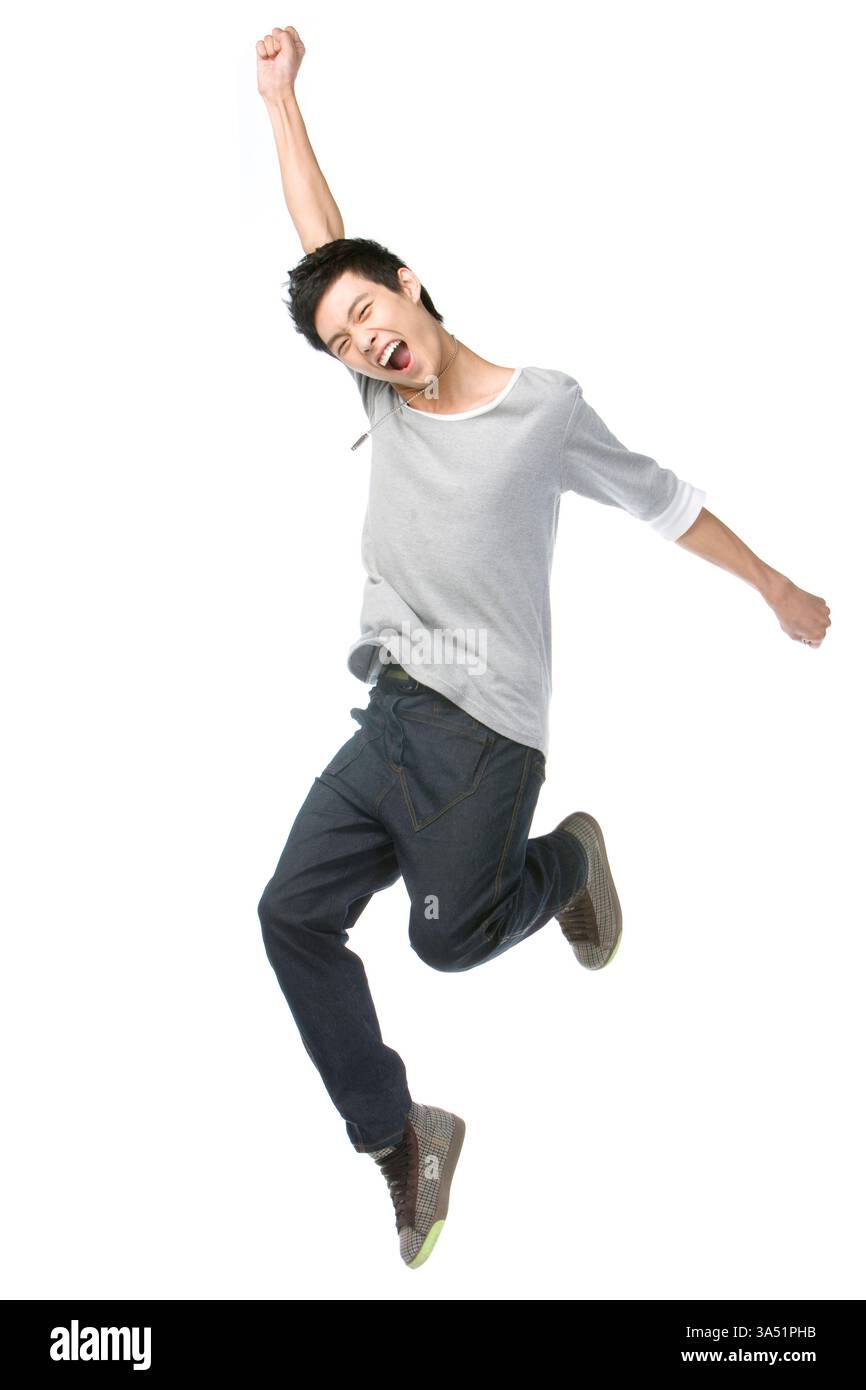 Jeune homme chinois saute en plein air les bras levés dans un studio sur fond blanc. Image de style de vie énergique pour la mode, la culture des jeunes et les campagnes de fitness avec une ambiance insouciante. Idéal pour la publicité et le matériel promotionnel ciblant les jeunes adultes. Banque D'Images