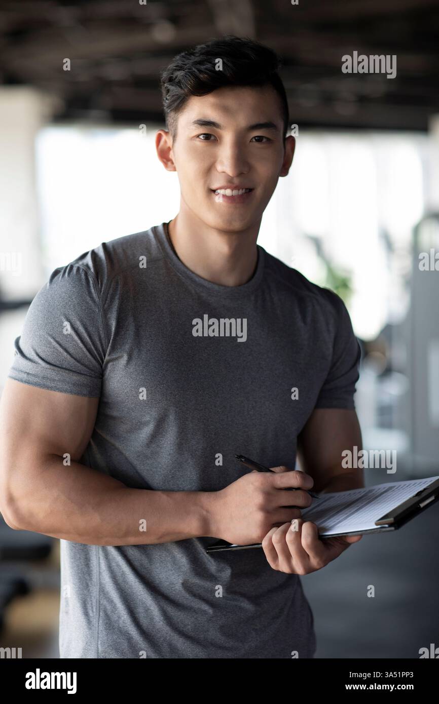 Un instructeur chinois de fitness souriant se tient debout dans une salle de gym, tenant un presse-papiers. Cette image de style portrait convient aux campagnes de coaching de fitness, de santé et de bien-être. Idéal pour les programmes de gymnastique, l'entraînement personnel et le contenu de bien-être. Banque D'Images
