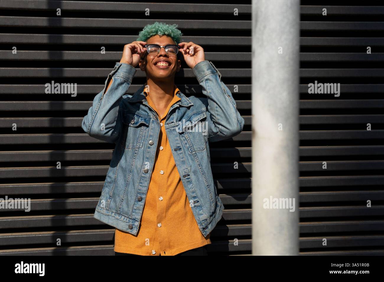 Homme hispanique tendance avec les cheveux teints en bleu, tenant des lunettes dans un look urbain décontracté. Une image audacieuse de mode et de style de vie adaptée au style de rue moderne, à l'expression personnelle et aux campagnes de lunettes. Idéal pour les articles sur la culture des jeunes et les tendances contemporaines. Banque D'Images