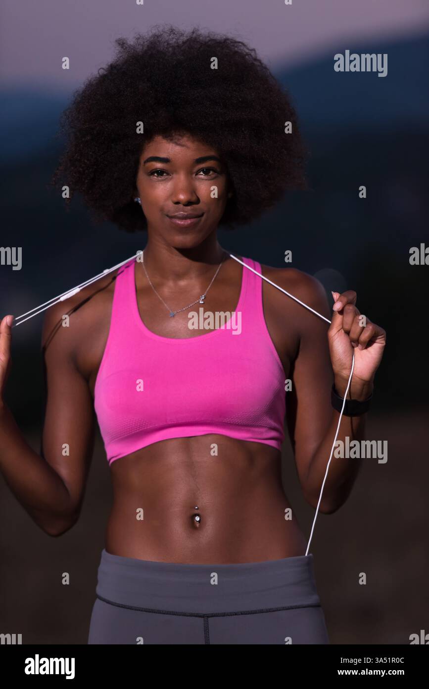 Portrait d'une jeune femme afro-américaine portant des écouteurs, faisant du jogging dans un pré la nuit pendant une nuit d'été. Ce moment de remise en forme en plein air capture la course à pied et un mode de vie sain dans la nature. Idéal pour les campagnes sur les femmes actives, l'entraînement cardio et les entraînements en plein air. Banque D'Images