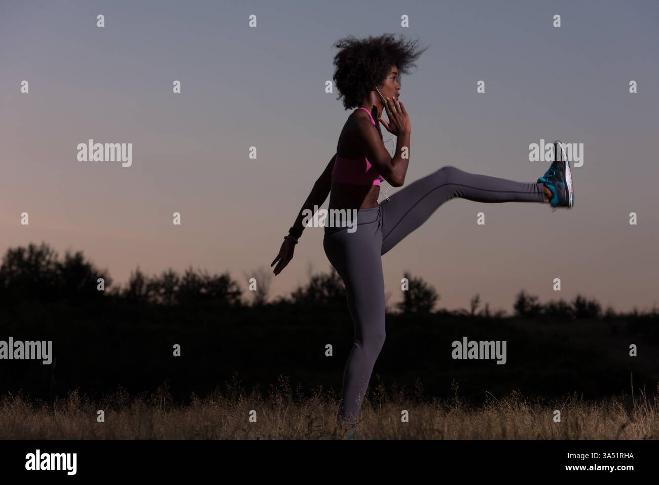 Jeune femme noire s'étire pour se détendre après un jogging dans un pré pendant une soirée d'été. Cette image de style de vie sportif met en valeur le fitness, le bien-être et les activités de plein air dans un cadre naturel. Idéal pour le sport, la santé et les campagnes de vie active. Banque D'Images